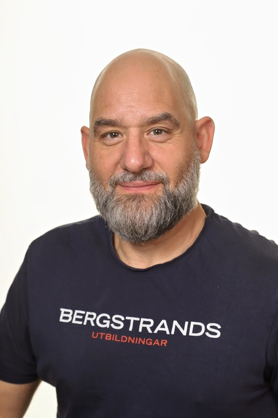 En skäggig man med flintskallig huvud är klädd i en marinblå T-shirt där det står "BERGSTRANDS UTBILDNINGAR" i vit och röd text. Han ler mot kameran mot en vanlig vit bakgrund.