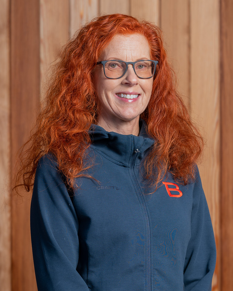 En kvinna med rött hår och glasögon som står framför en trävägg.
