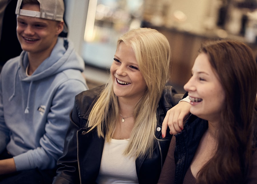 Bergstrands elever Tre personer som sitter tillsammans, ler och skrattar. Personen till vänster bär en blå luvtröja och keps, personen i mitten bär en skinnjacka och personen till höger bär en brun topp.