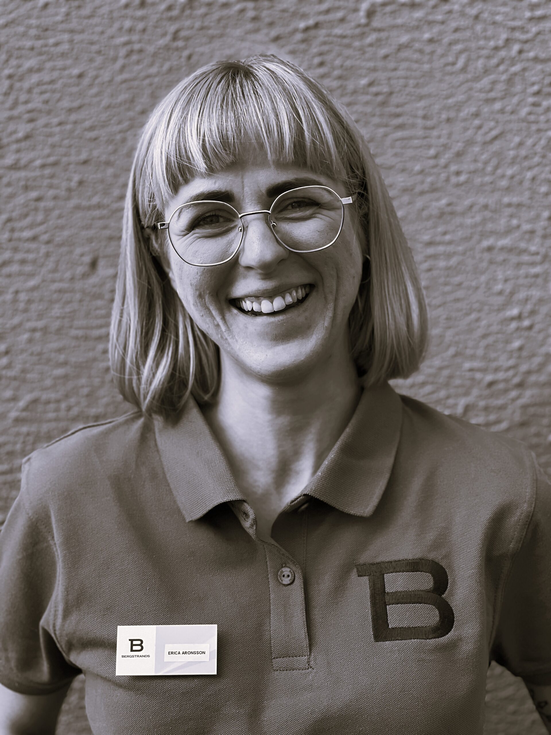 En person med glasögon och rakt ljust hår ler mot kameran, klädd i en skjorta med krage och en namnskylt med bokstaven "B" på både skjortan och skylten.
