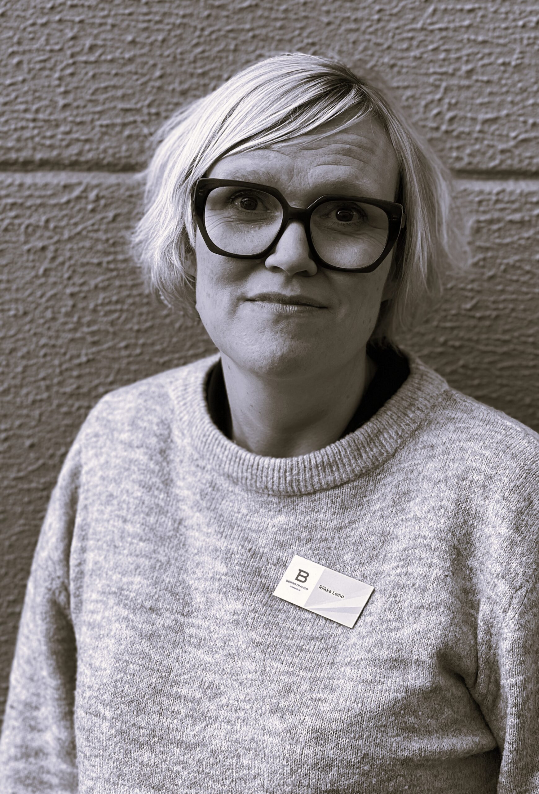 Person med kort ljust hår och glasögon, iklädd tröja och namnskylt, står framför en strukturerad vägg.
